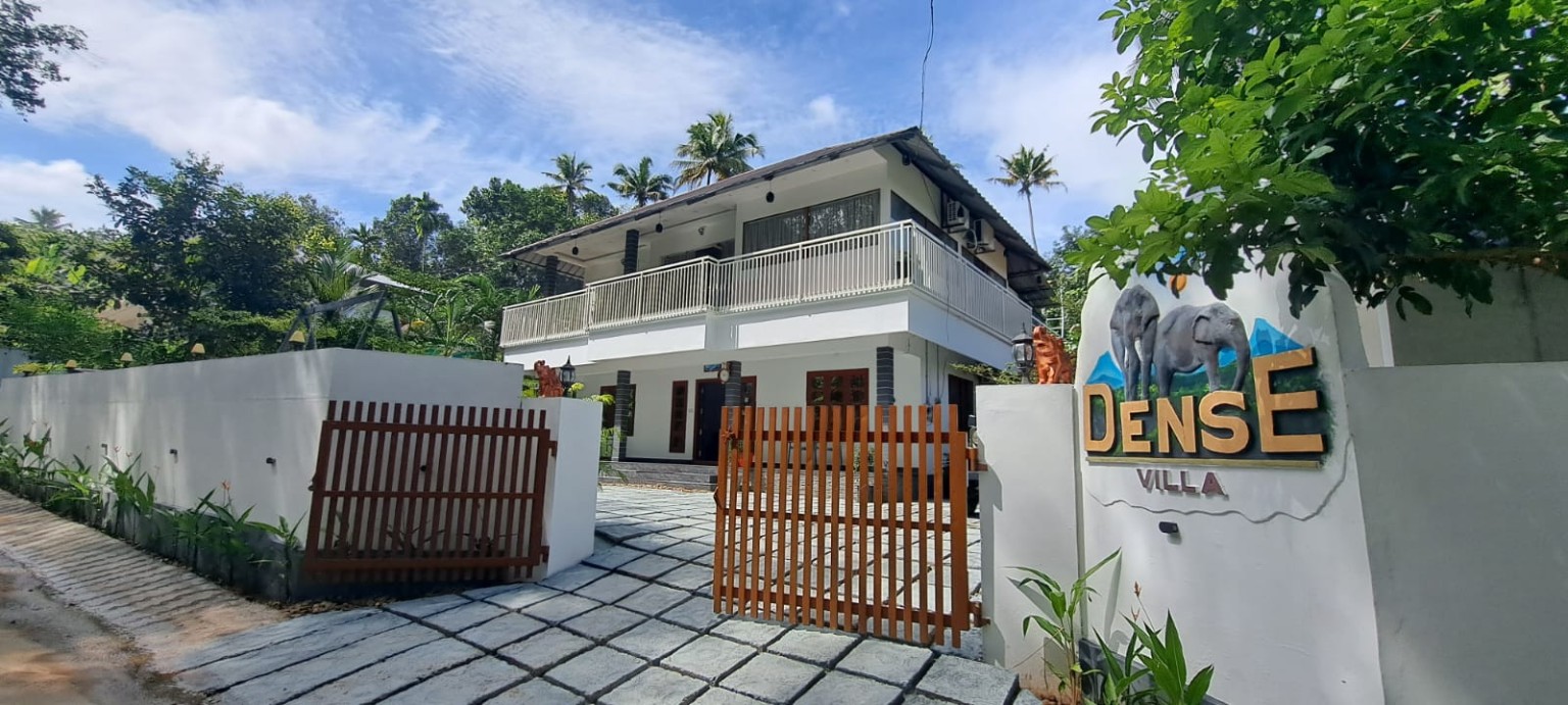 Dense Villa at Kuttampuzha seen from the lane: a white two-storey villa with a long first-floor balcony, timber gates and coconut palms behind
