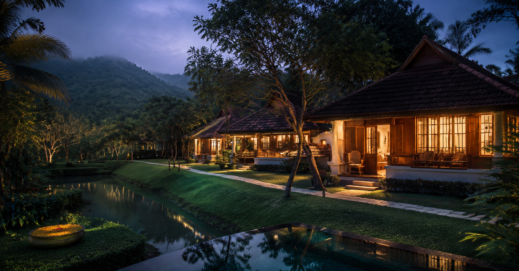 Illustrative cinematic view of a tiled-roof Kerala-style villa lit at dusk beside a still water channel, with misty forested hills behind