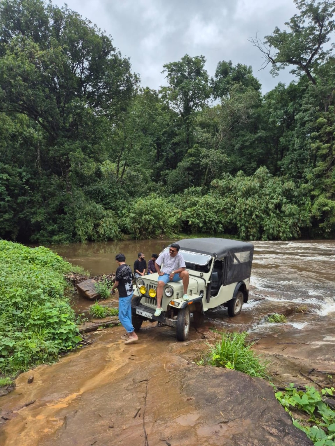 A jeep on a wet rock shelf beside a fast monsoon river at Kuttampuzha, dense forest on the far bank
