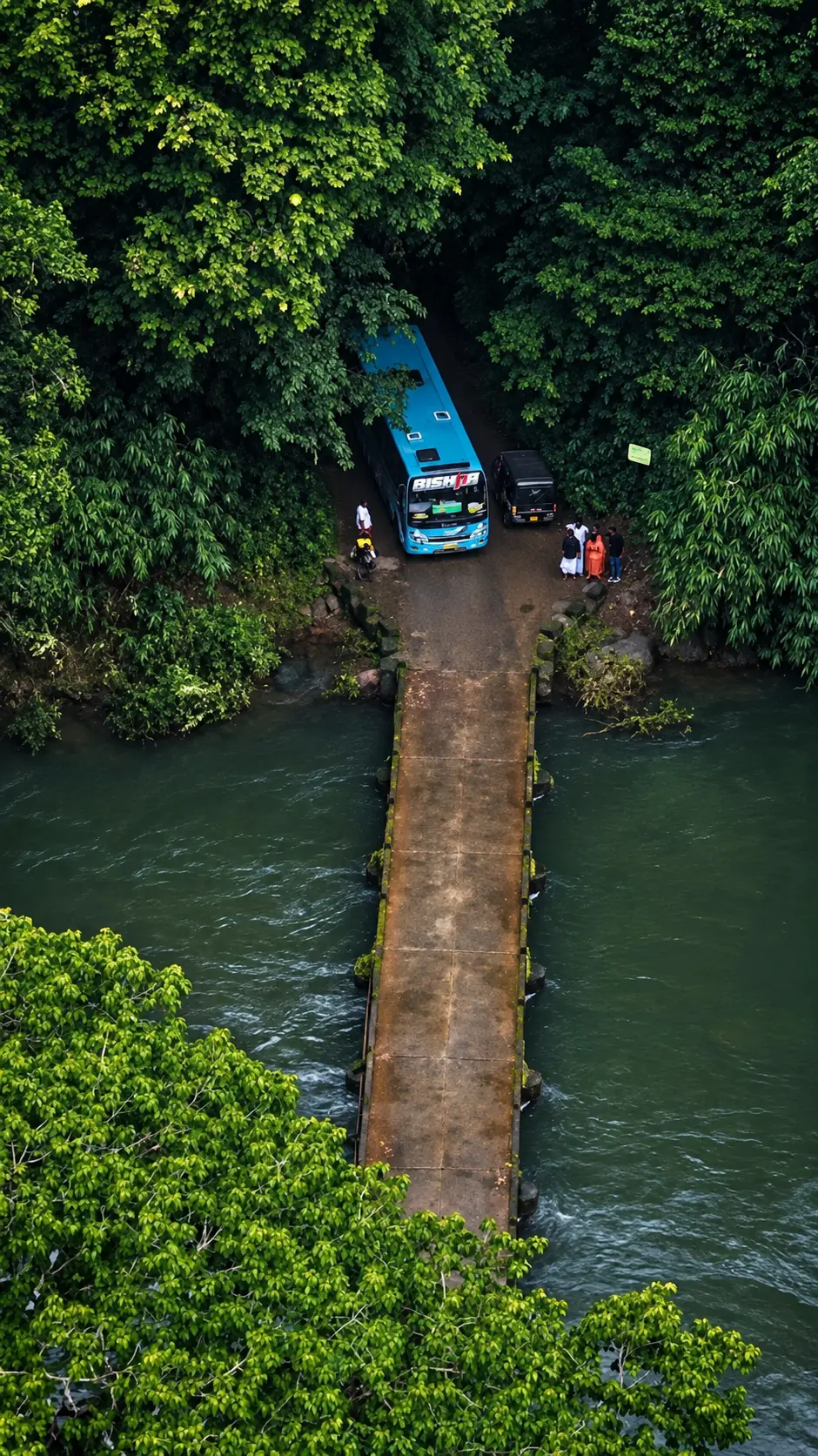 Aerial view of a narrow concrete bridge crossing a green river, with a bus waiting on the forest bank