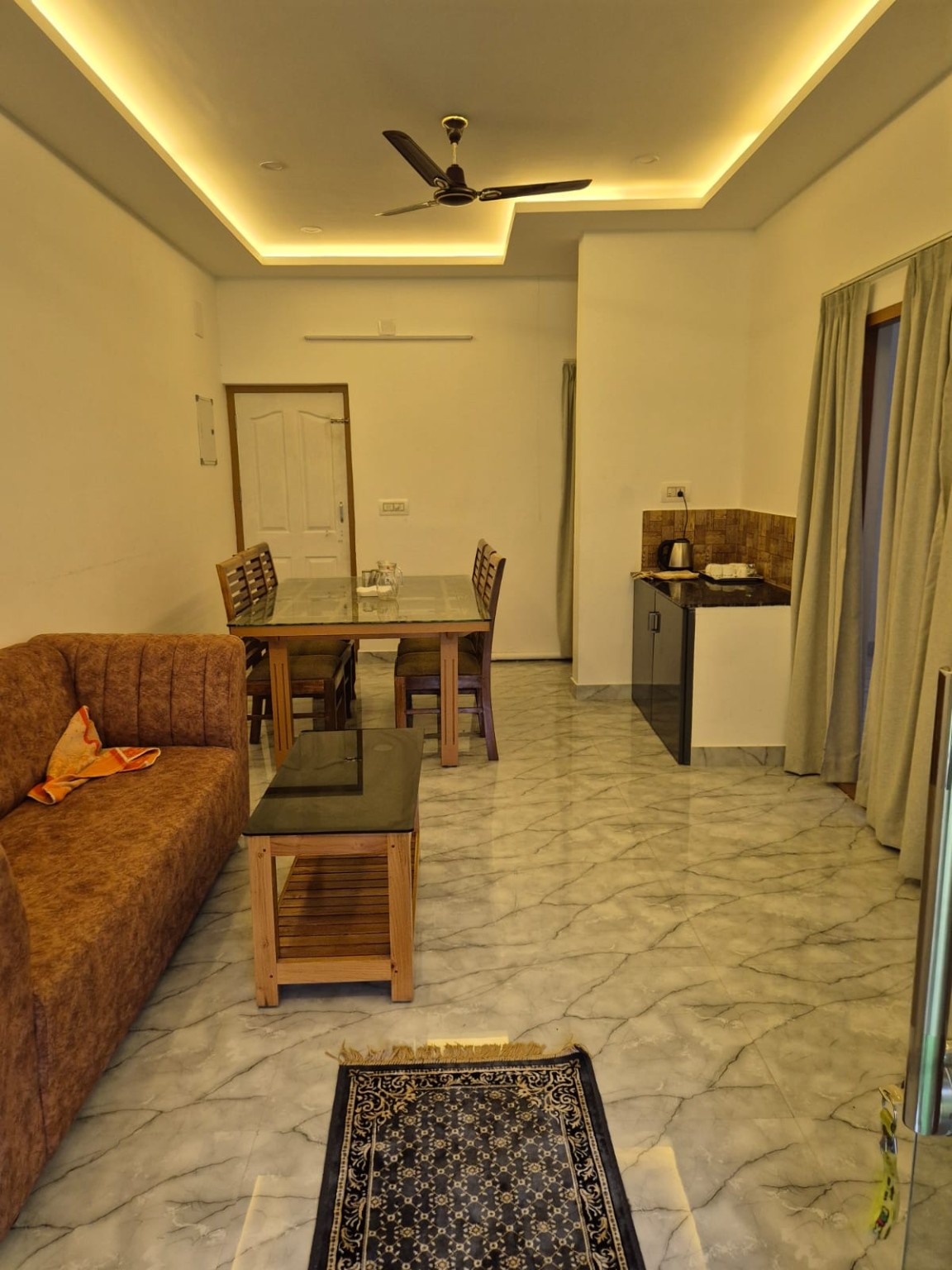Living and dining room at Dense Villa with a sofa, glass-topped dining table for four, coffee table and kitchenette counter under warm cove lighting